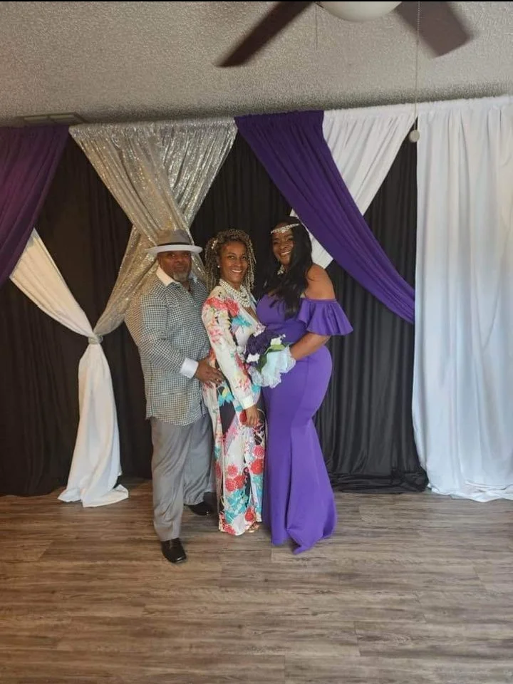 Three people standing in front of a decorative backdrop with purple, black, white, and gold drapes, dressed in formal attire, smiling for a photo.