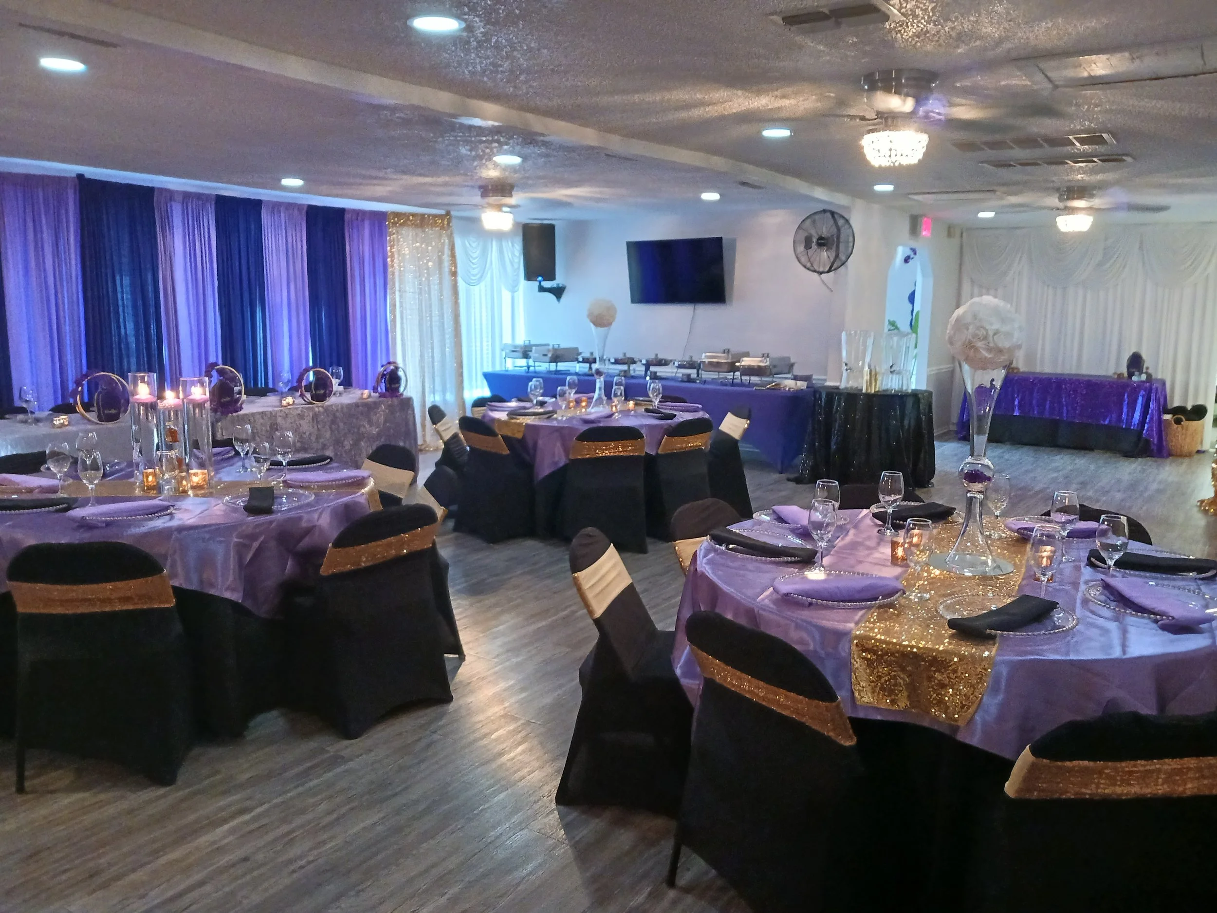Indoor banquet setup at A Level Up Rental Hall in Tampa with purple linens, black chair covers, gold accents, candles, and floral centerpieces.