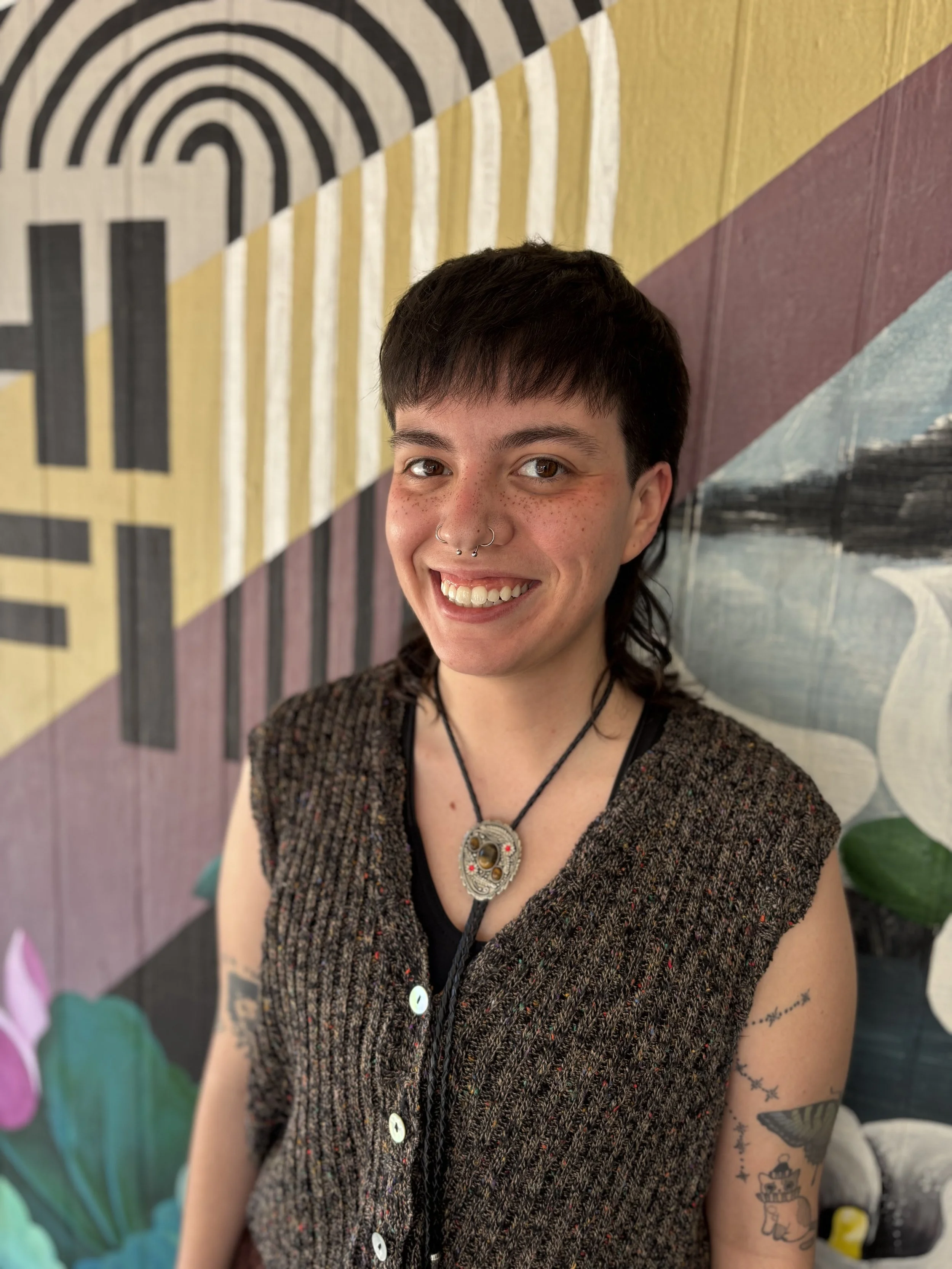 A young woman with short dark hair, a nose ring, and tattoos on her arm, smiling in front of a colorful mural background.