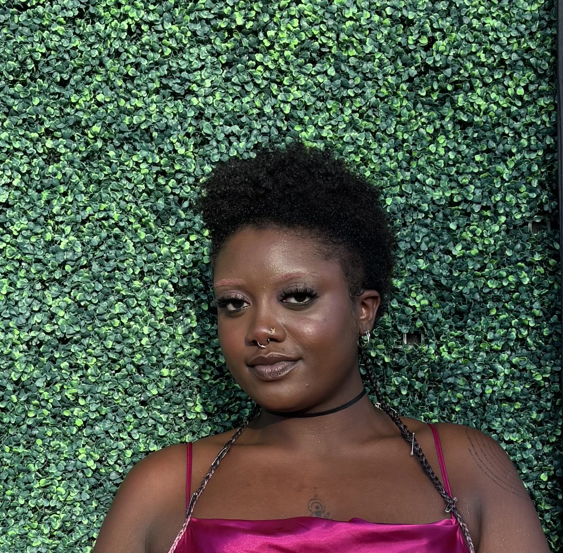 Portrait of a young Black woman with natural hair, wearing makeup, a septum piercing, earrings, a black choker, and a pink satin top, standing in front of a green leaf wall.
