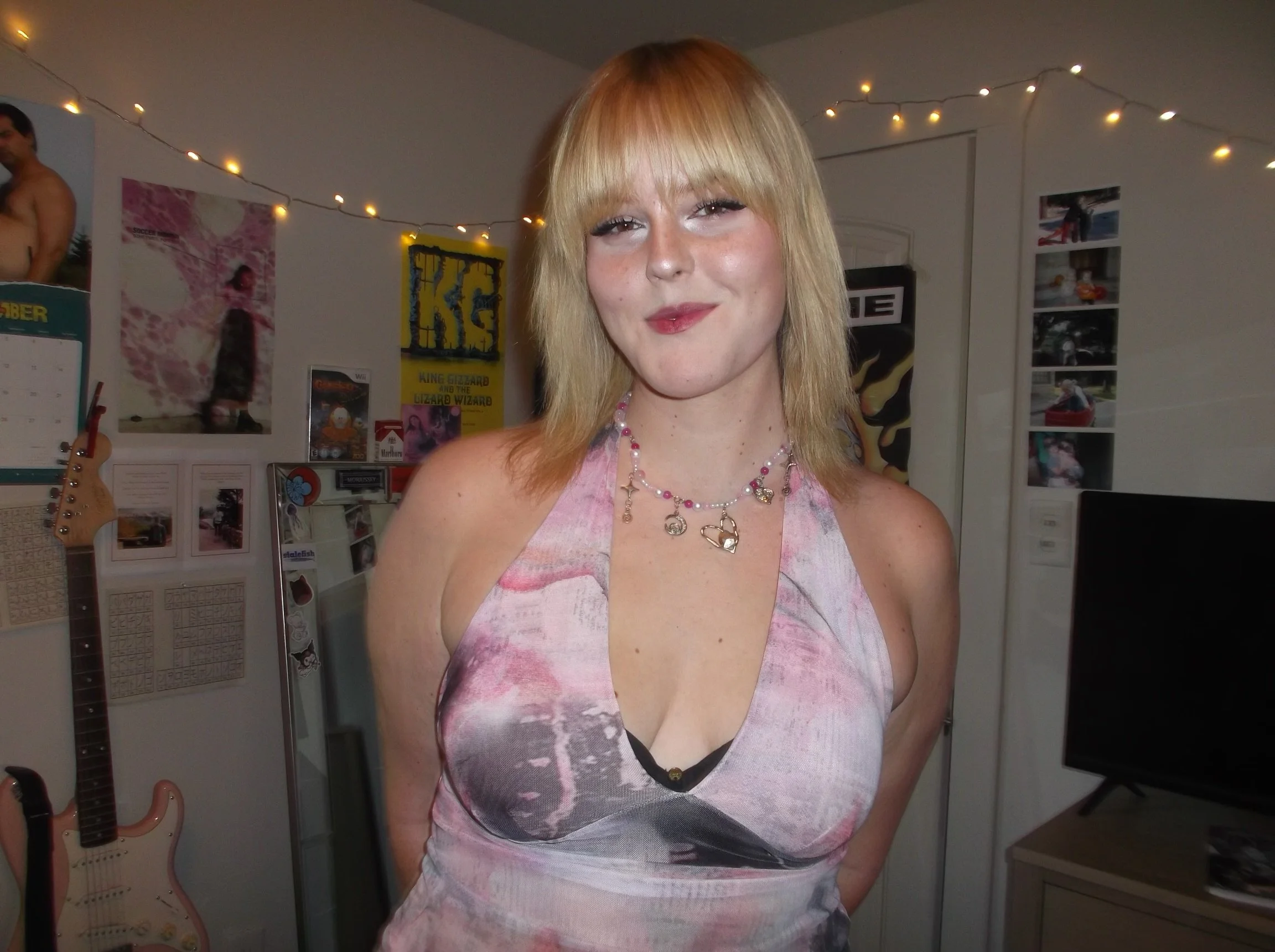 A young woman with blonde hair and a pink and gray tie-dye dress standing in a room decorated with posters and string lights.