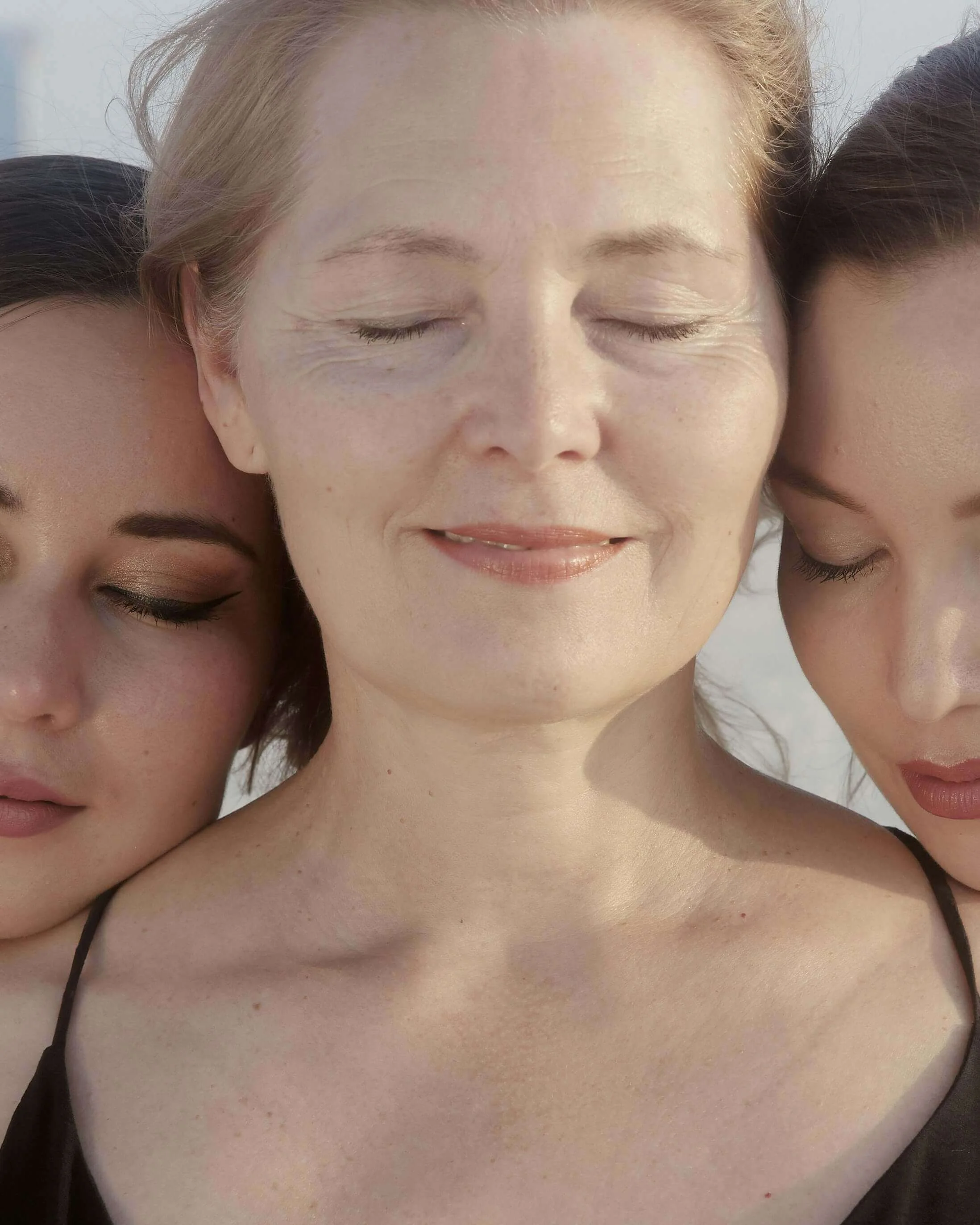 Three women with closed eyes and smiling faces, close-up of their faces and shoulders, some wearing black clothing, with light skin and different hair colors.