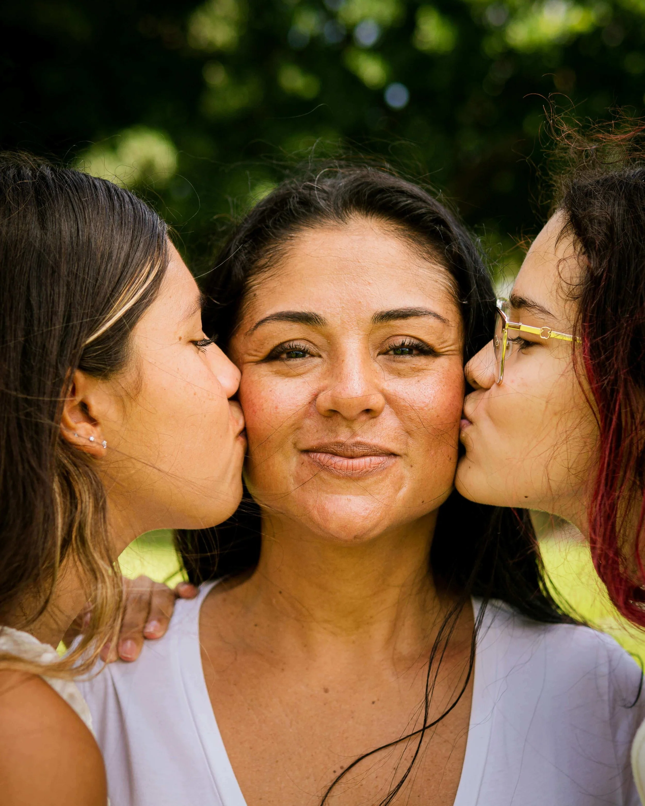 A woman wearing a white top being kissed on each cheek by two young girls, one on each side, in an outdoor setting with green foliage in the background.