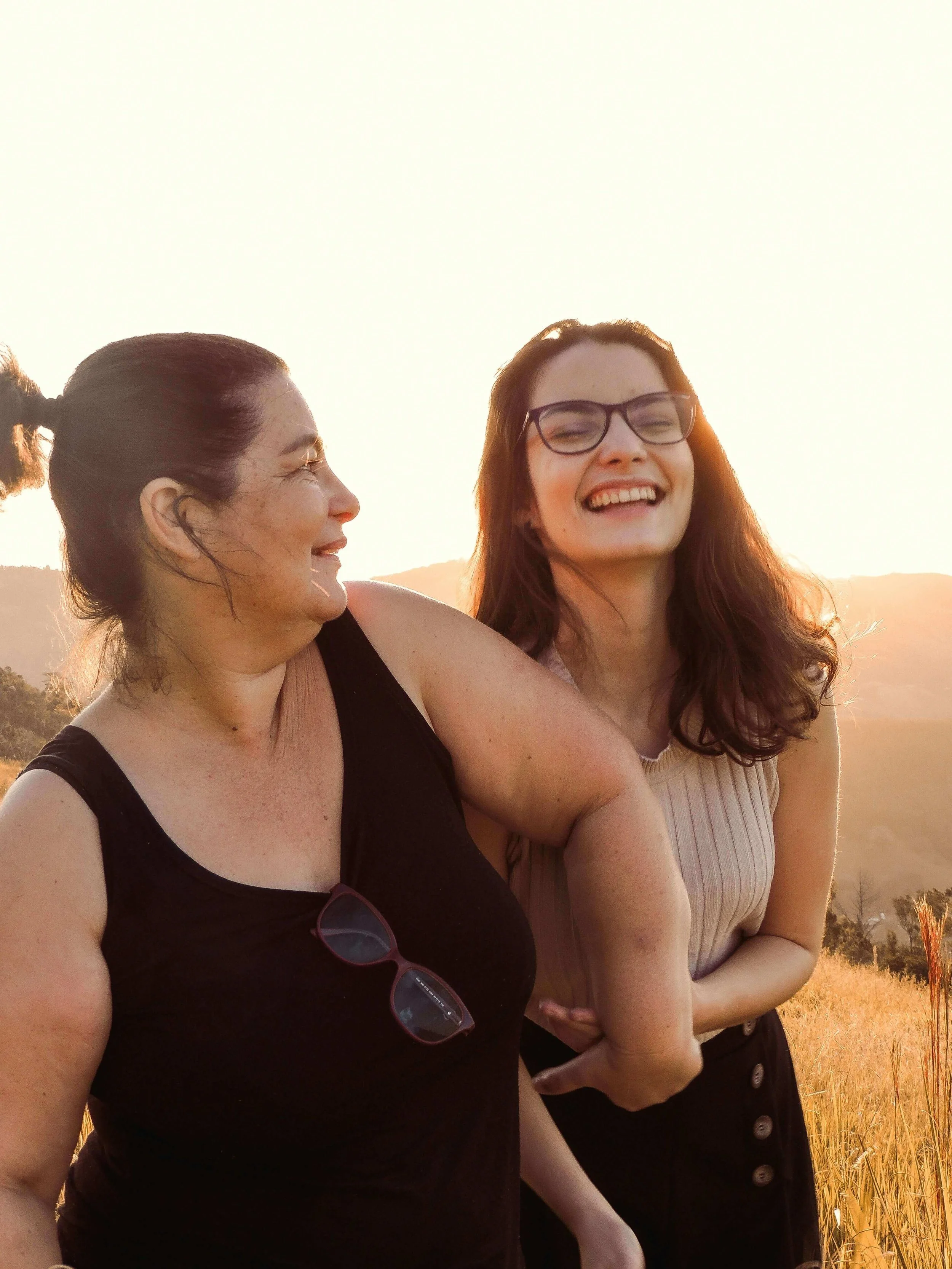 Two women smiling and talking outdoors during sunset, standing in a field of tall grass with mountainous landscape in the background.