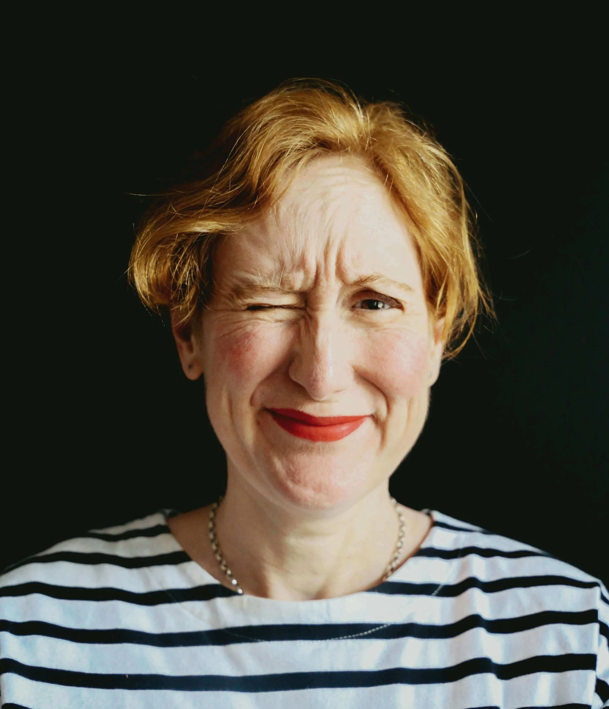 Close-up of a woman with fair skin, short reddish hair, red lipstick, and a striped shirt, smiling and winking against a black background.