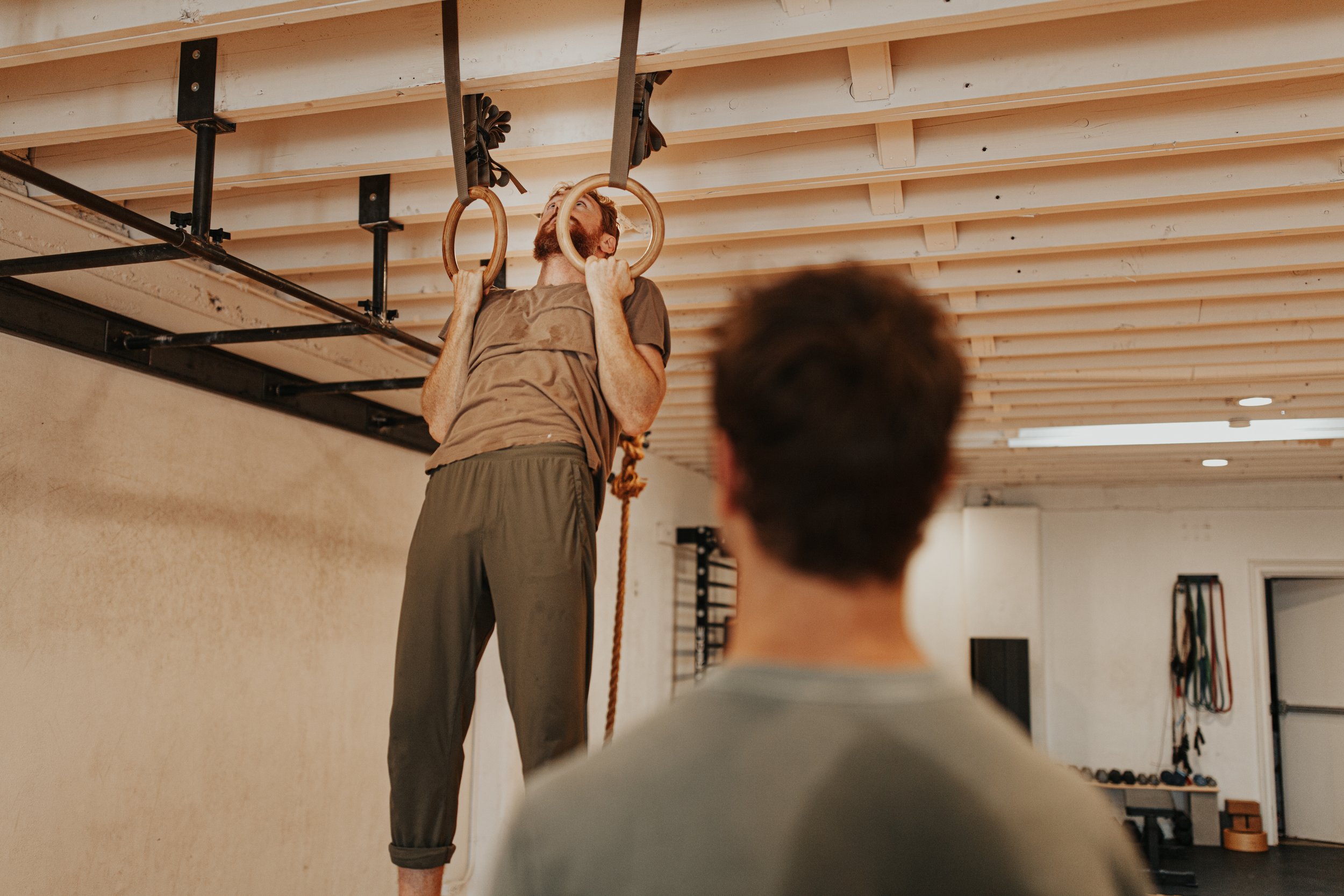 A man with a beard is hanging from gymnastic rings attached to the ceiling in a fitness studio. A trainer or instructor is standing nearby, possibly coaching. A person with a short haircut and gray shirt is sitting on the floor in the foreground, observing the activity.