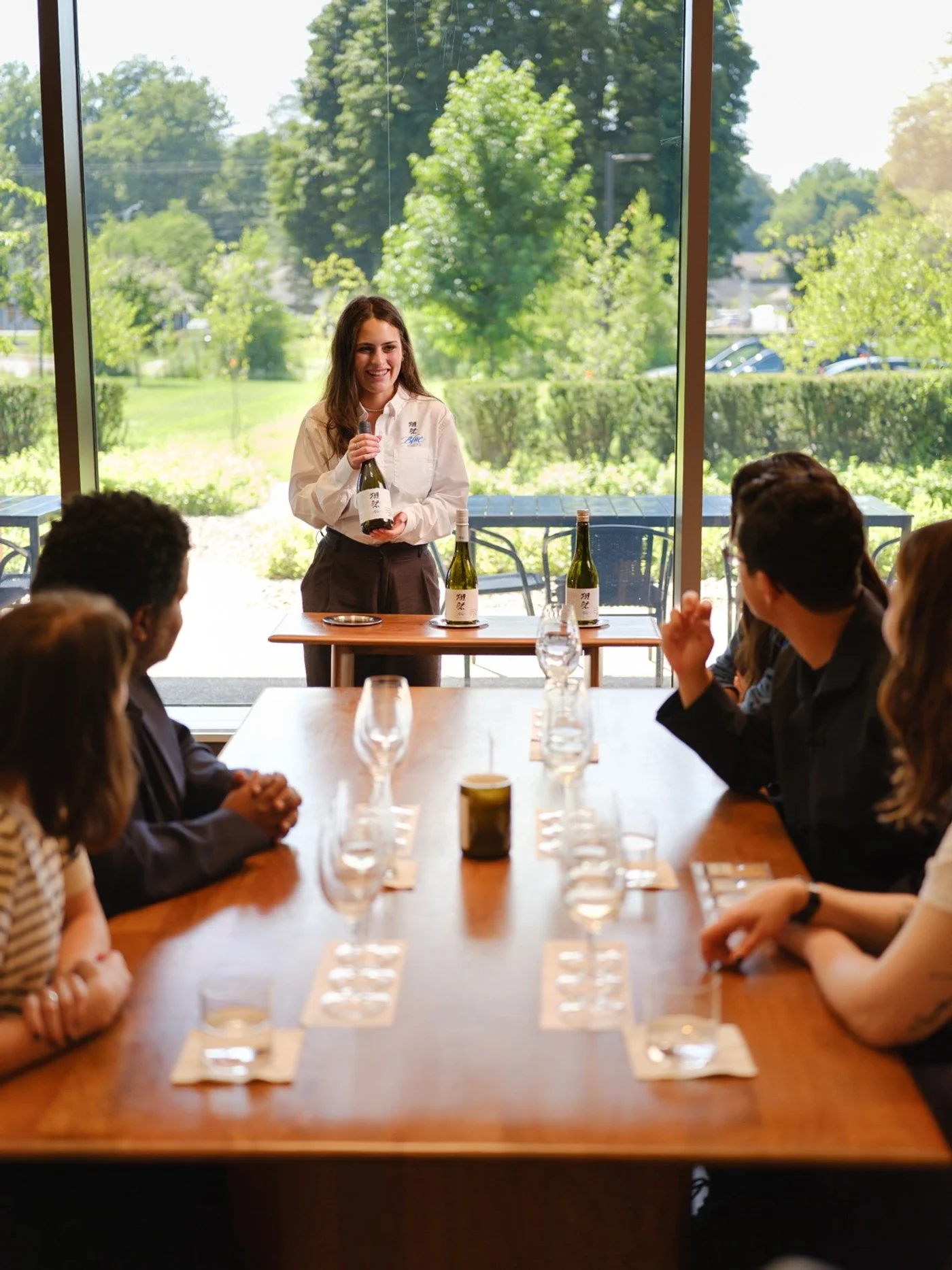 woman-presenting-wine-to-a-seated-group-in-a-bright-room-with-large-windows.jpg.webp