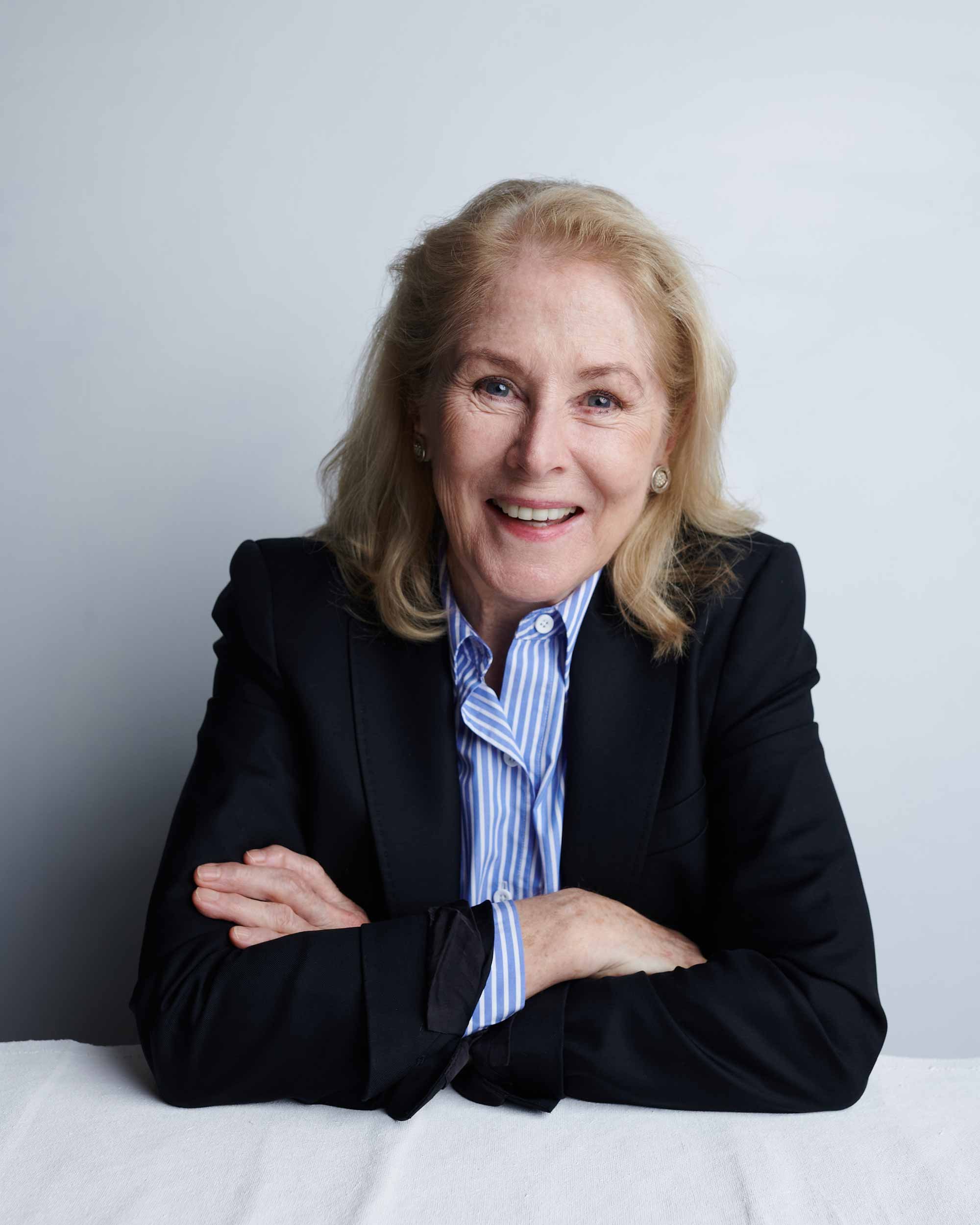 A woman sits a a desk with her arms folded on the table smiling at the camera