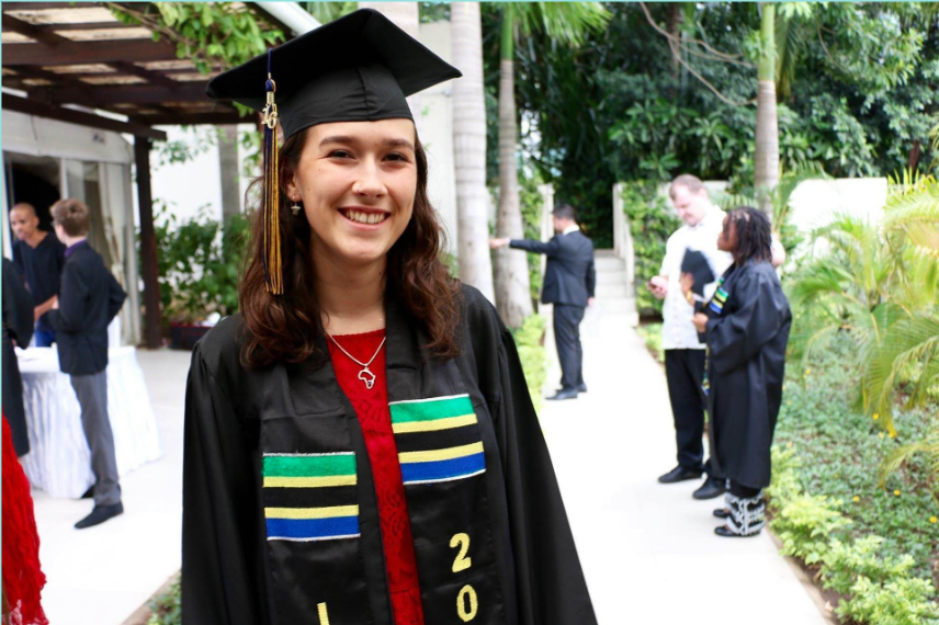 Young woman in graduation gown and cap smiling at camera at outdoor graduation ceremony with other graduates in the background.