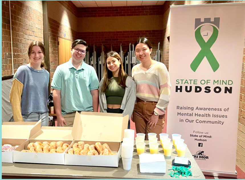 Four young adults standing behind a table with food boxes, cups, and promotional materials at a mental health awareness event, with a banner reading "State of Mind Hudson" in the background.