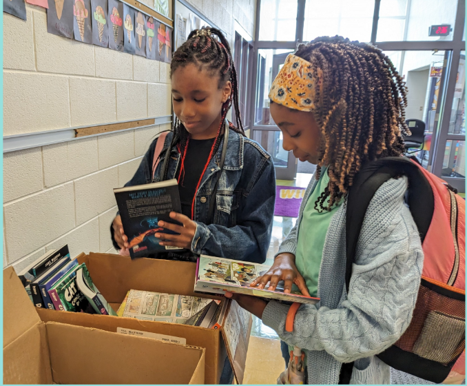 Two girls look through books at a school book sale, with boxes of books on a table in a school hallway.