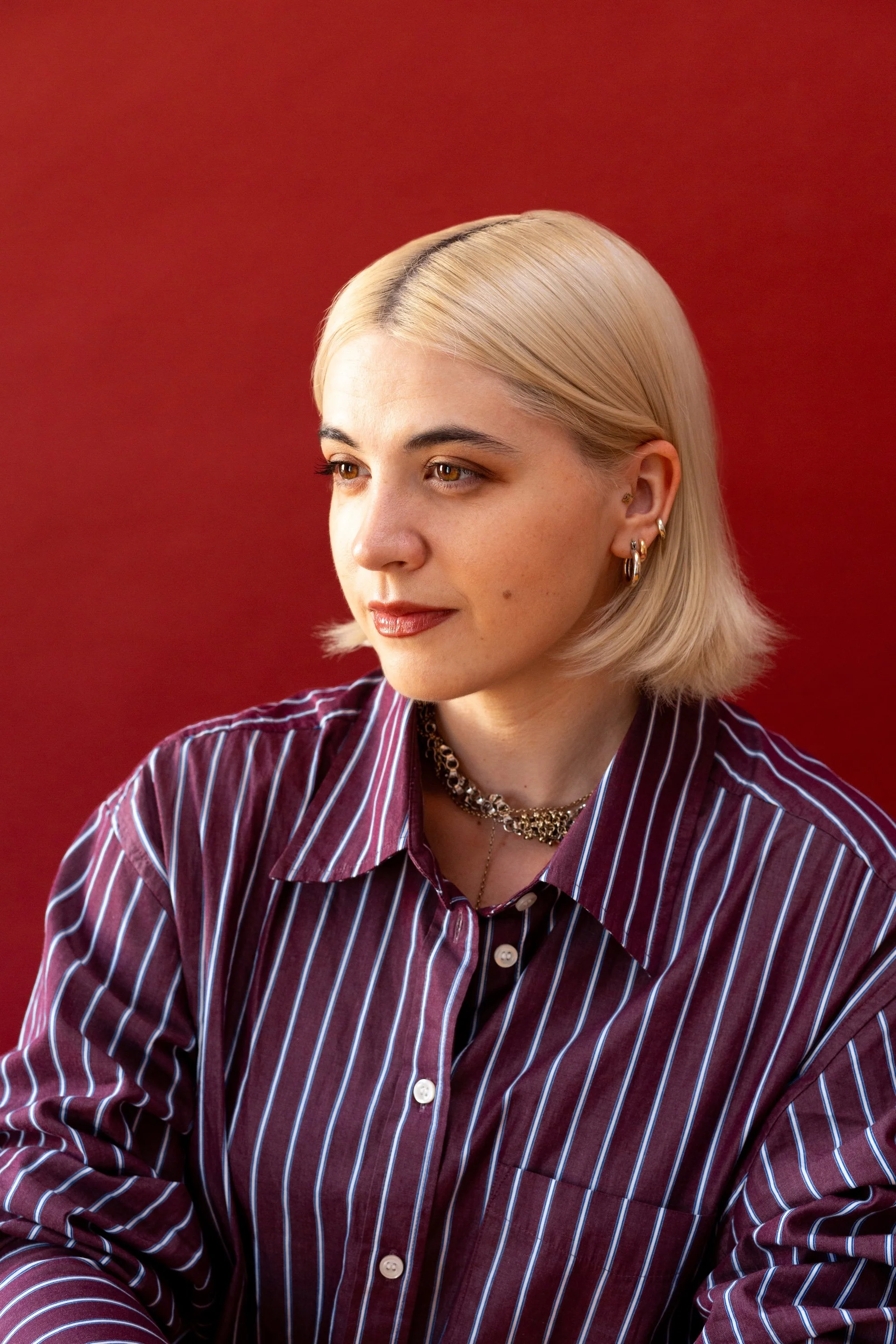 Photo of Gabriela looking off to the side with short blond hair in a maroon, blue, and white striped button down, on a deep rich oxblood red background.