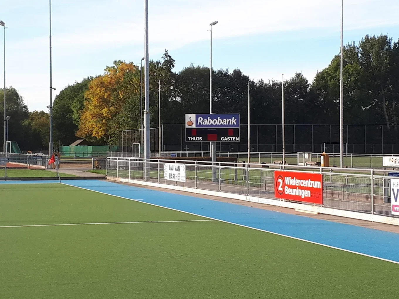 An empty outdoor sports field with green and blue turf, a scoreboard, and advertising banners, surrounded by trees and tall stadium lights.