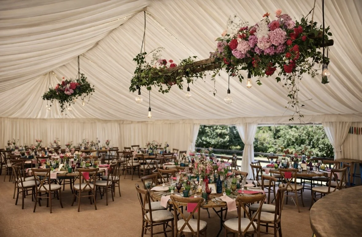 Decorated event tent with round tables set for a celebration, featuring floral centerpieces and hanging pink and purple hydrangea arrangements with Edison bulb string lights.