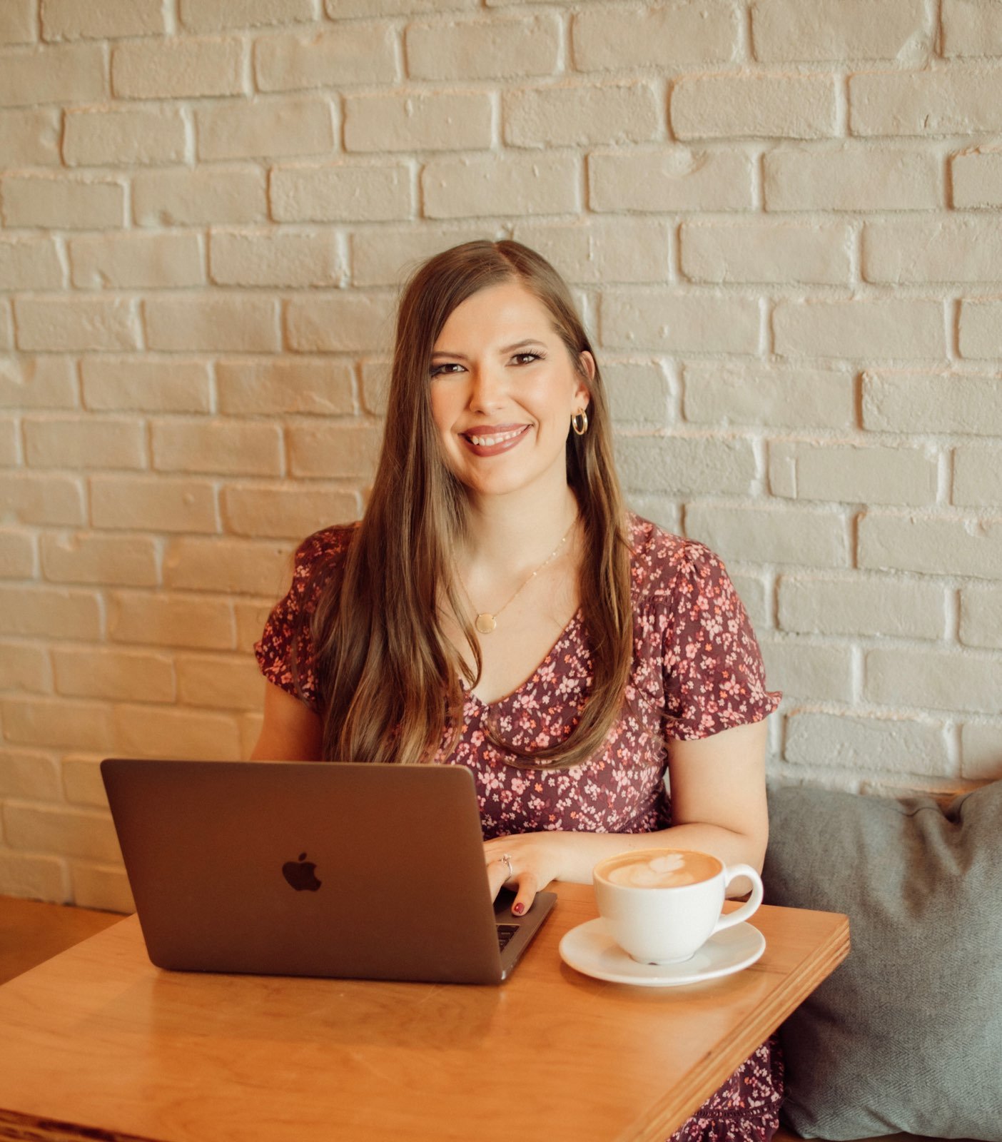 A woman with long brown hair and a floral dress sitting at a wooden table in a cafe, smiling at the camera with a laptop and a cup of coffee in front of her.