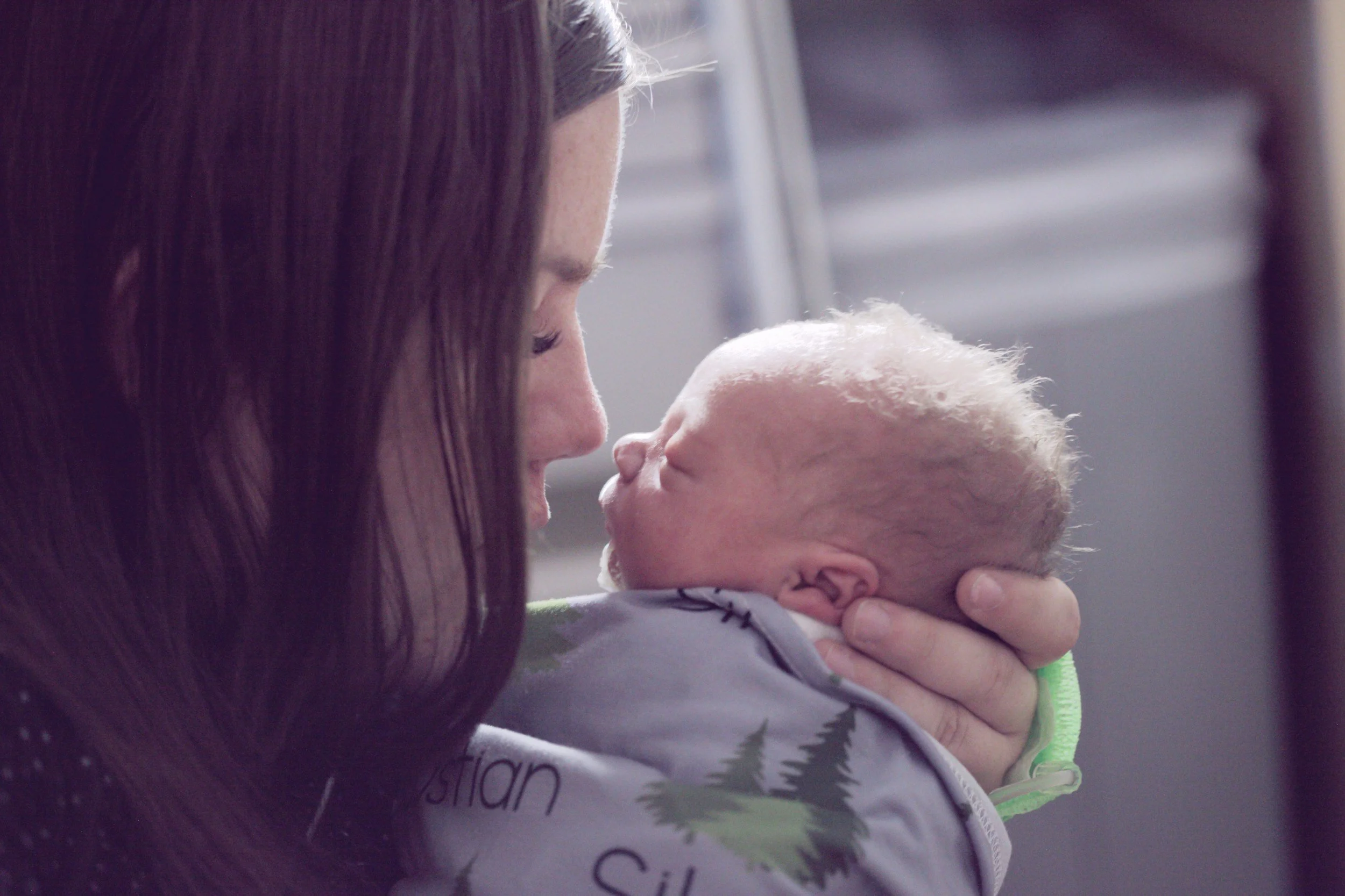 A woman holds a newborn baby close to her face, both looking at each other with affection. The woman has long brown hair, and the baby has light hair and fair skin. The background is blurred, focusing on the emotional interaction.