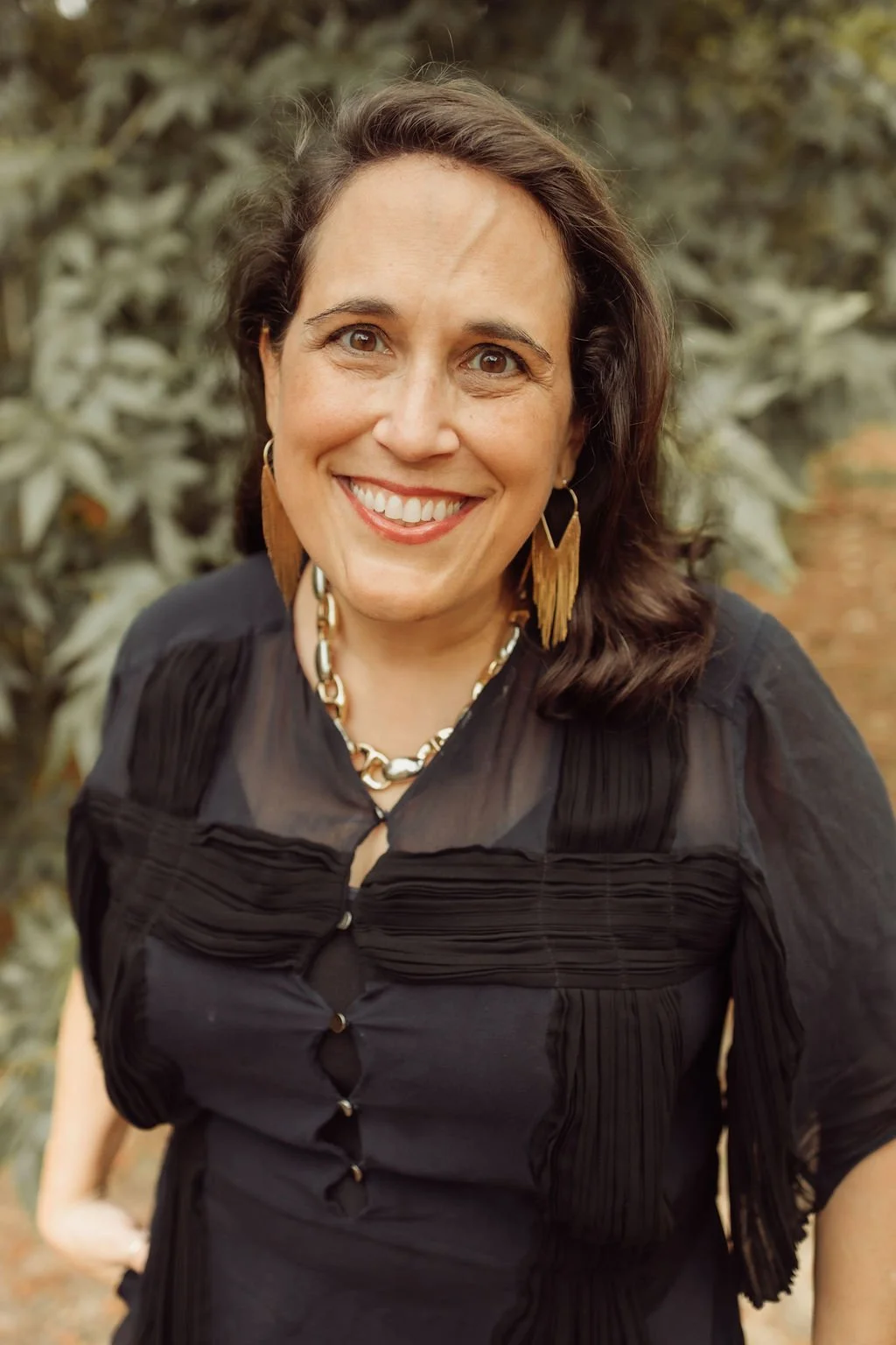 A portrait of a woman with shoulder-length curly brown hair, smiling, wearing makeup, a black top, gold jewelry, and a green and black blurred background.