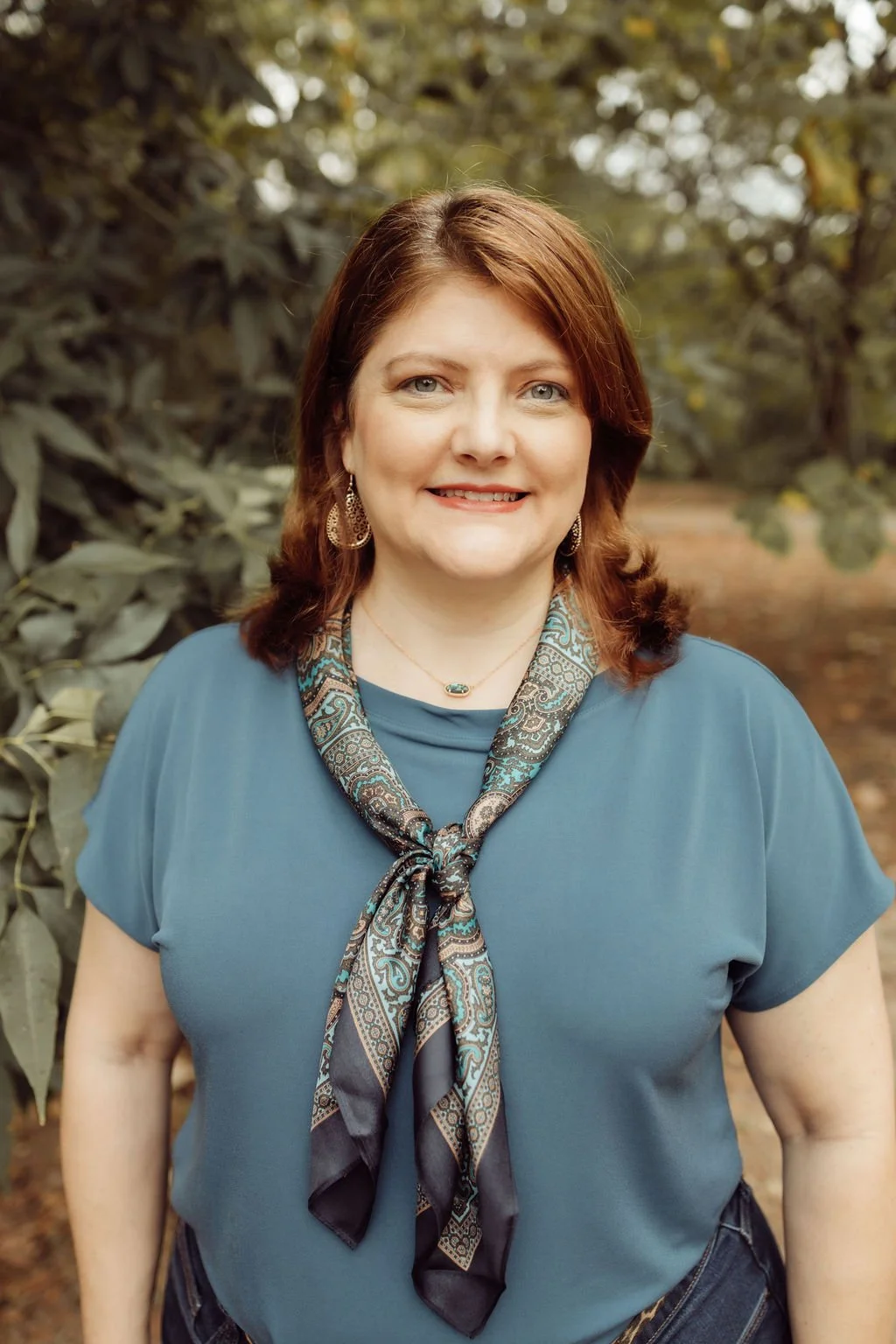 A woman with shoulder-length brown hair, wearing a blue top and blue glasses, smiling at the camera in front of beige curtains with a green plant nearby.