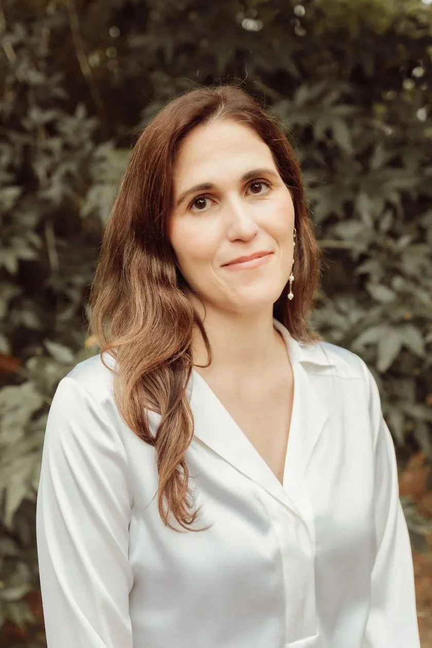A woman with long wavy brown hair, wearing a white blouse, standing outdoors in front of green foliage.
