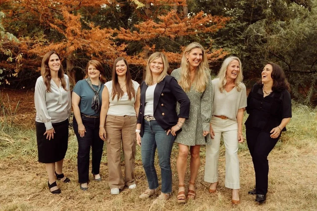 Group of seven women standing outdoors in front of fall-colored trees, smiling and laughing.