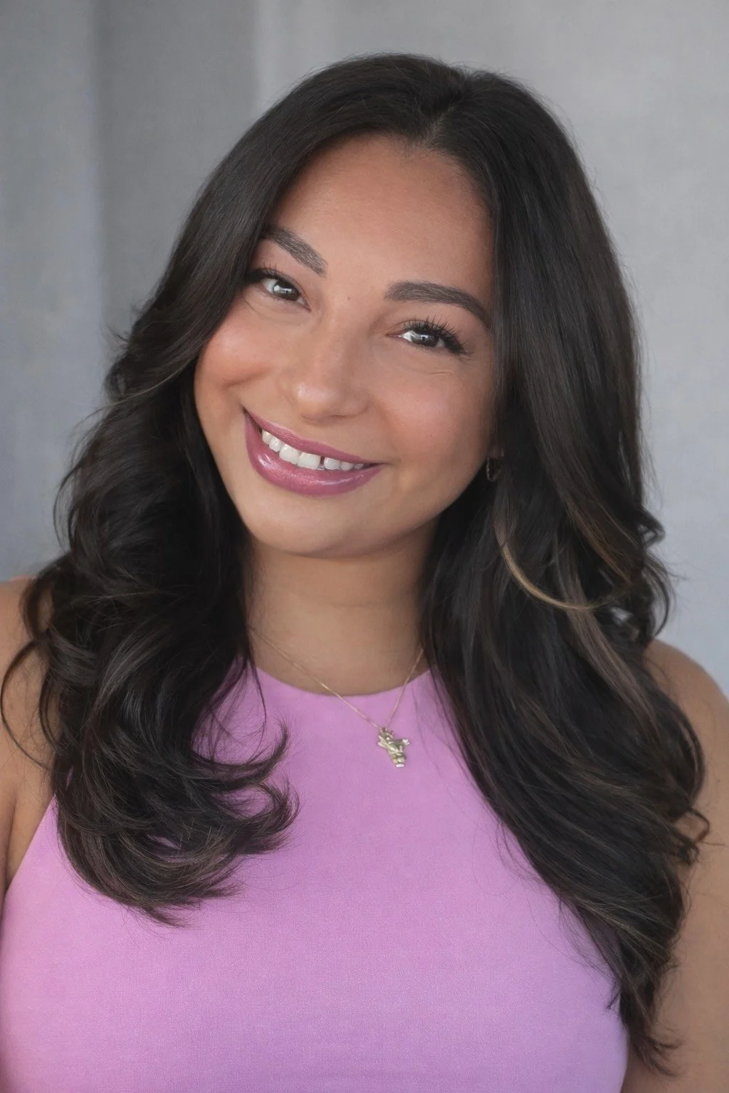 Portrait of a smiling woman with long dark wavy hair wearing a purple top and a gold necklace.