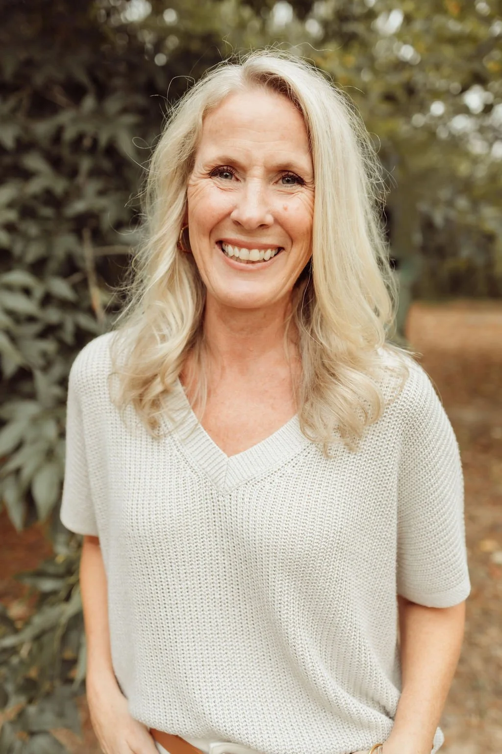 A woman with long blond hair smiling outdoors with trees and foliage in the background.