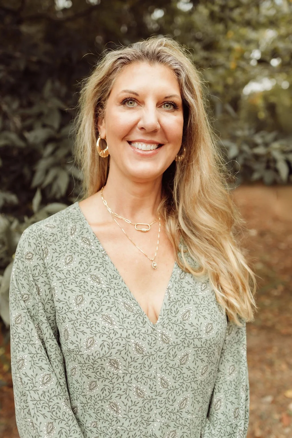 A woman with curly blonde hair smiling outdoors, wearing a striped shirt and layered necklaces.