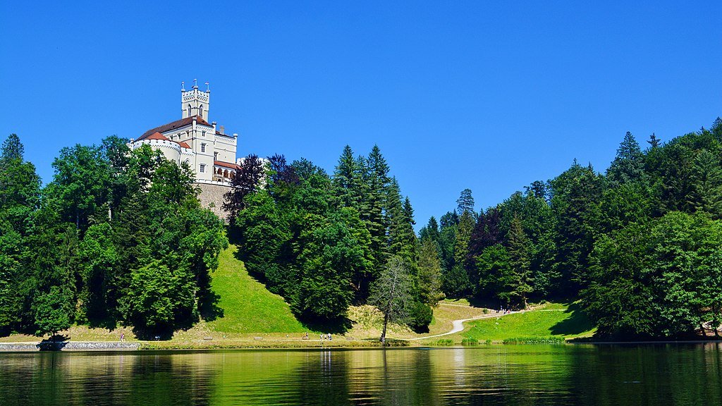 Trakšoćan Castle sits on a hill surrounded by green pine trees as seen from the waters of a pond below.