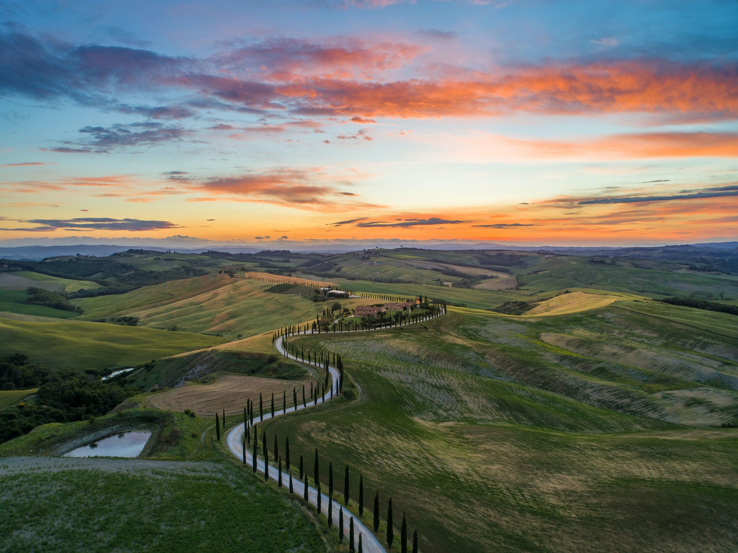 An aerial view over a winding dirt road in the countryside of Tuscany, with gently sloping green hills all around, and a magnificent sunset