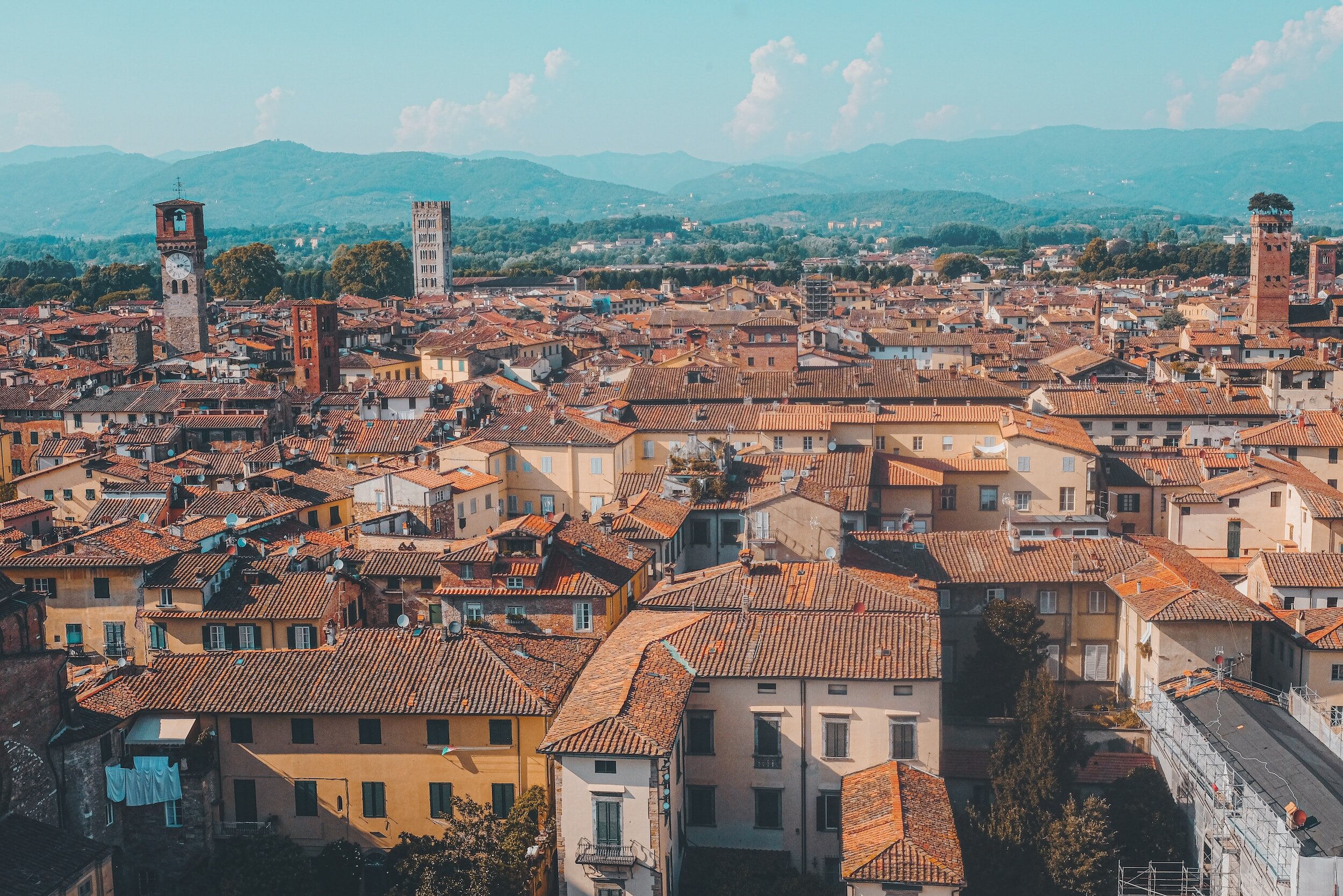 An aerial view over the red roofs of the city of Lucca, with tree-covered hills in the distant background