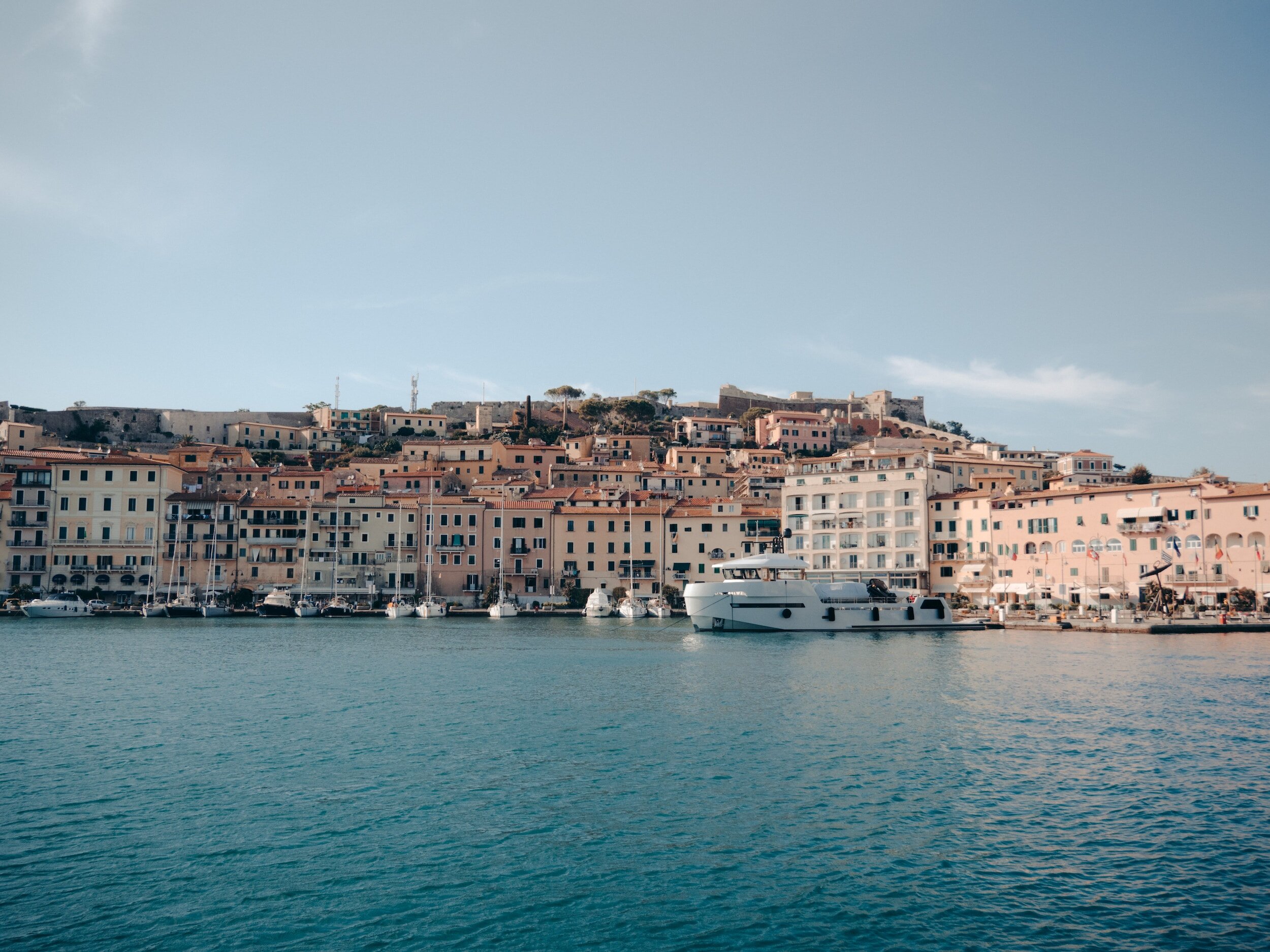 The waterfront of the town of Portoferraio, lined with buildings rising up the surrounding hill, seen from the water