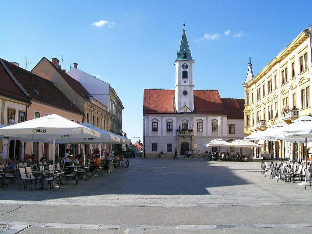 A city square lined with outdoor tables under white umbrellas in the town of Varaždin, Croatia.