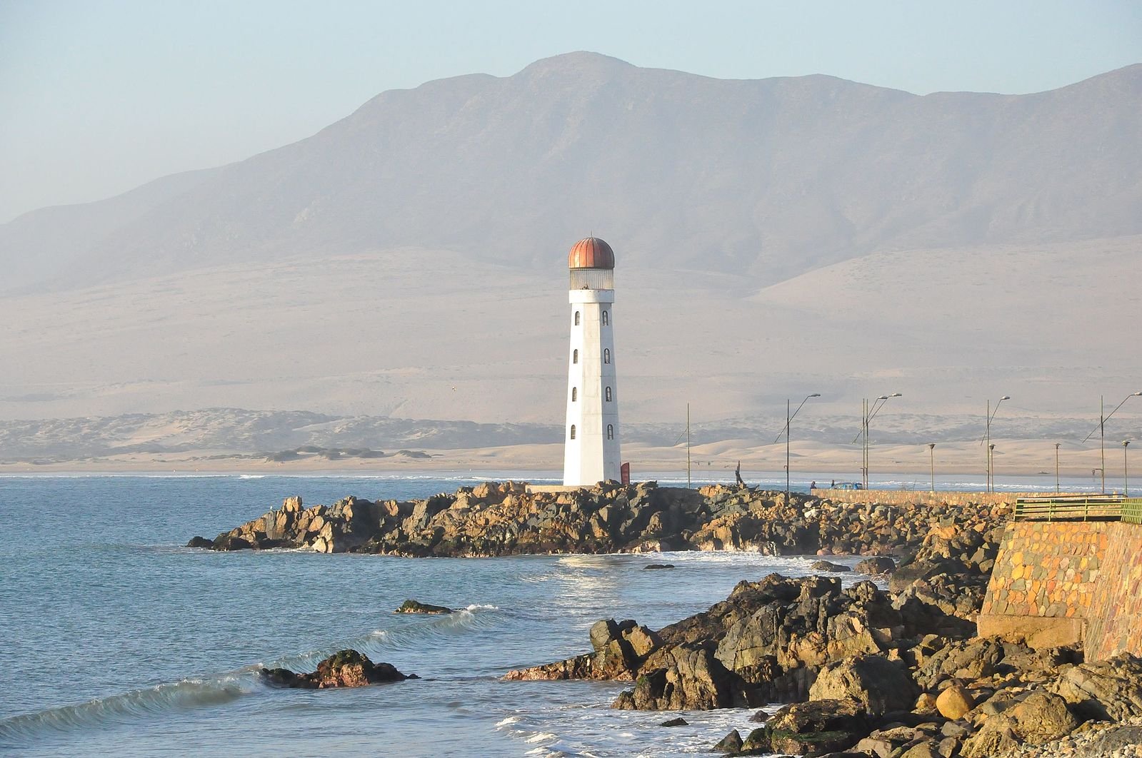Huasco's Faro Monumental lighthouse on a rocky outcrop surrounded by calm sea with high desert mountains hazy in the distance.