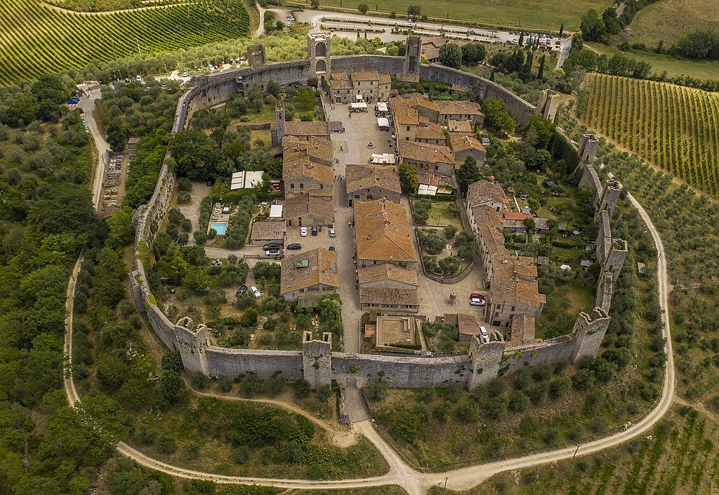 An aerial view of the town of Monteriggioni, with its medieval walls encircling the town's buildings, and surrounded by trees and fields of grapes around it