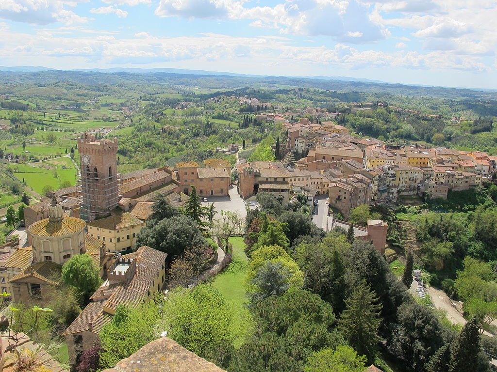 An aerial view of the town of San Miniato, with its medieval buildings clustered around one another, and green fields all around
