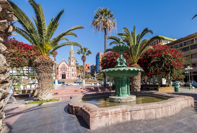 A fountain, palm trees, and colorful flowers dot the square in front of San Marcos Cathedral in Arica, Chile.