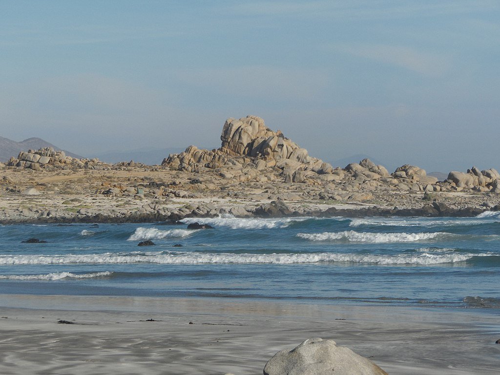 Small waves break on the coast with large boulder formations visible beyond them on a beach in Llanos del Challe National Park.