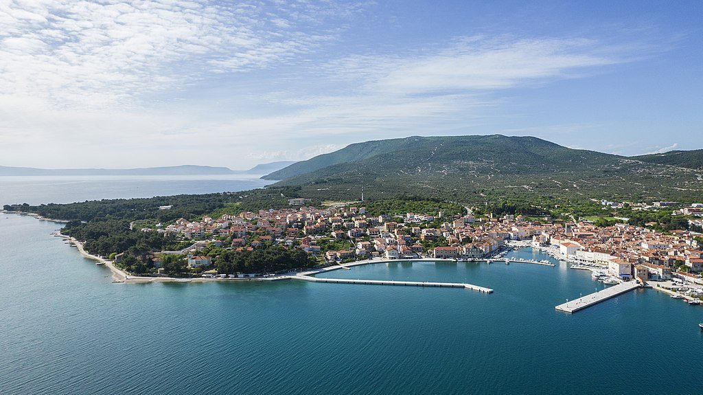 An aerial view of the town of Cres with its protected marina in the water and a green valley behind it.