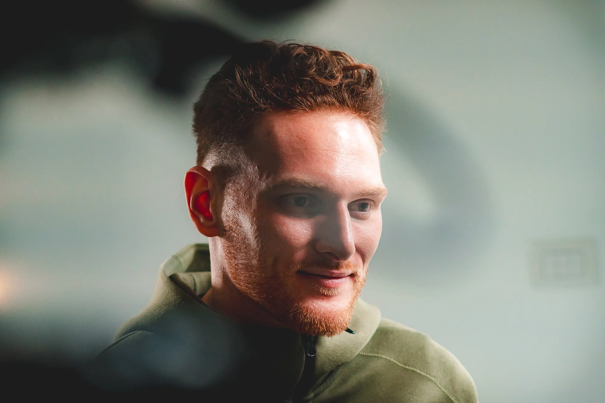 A young man wearing a green hoodie, looking down with a slight smile, being interviewed on a research project.