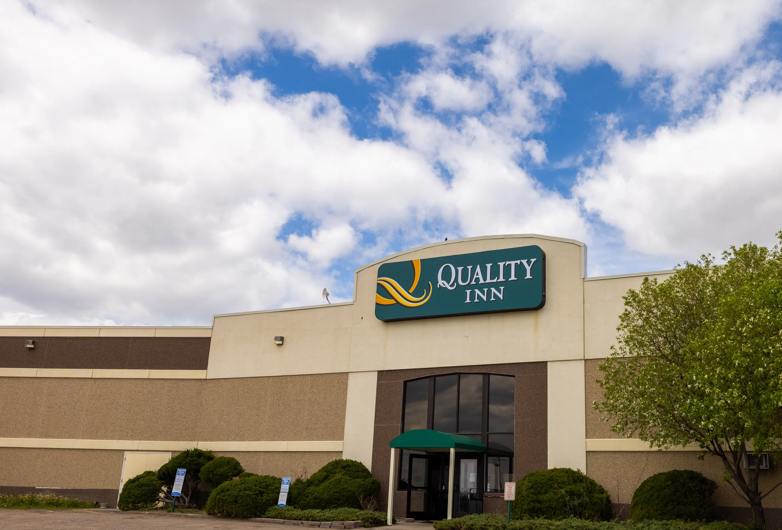 Exterior of a hotel named 'Quality Inn' with a large sign, a green awning over the entrance, and surrounded by greenery and parking signs, under a partly cloudy sky.