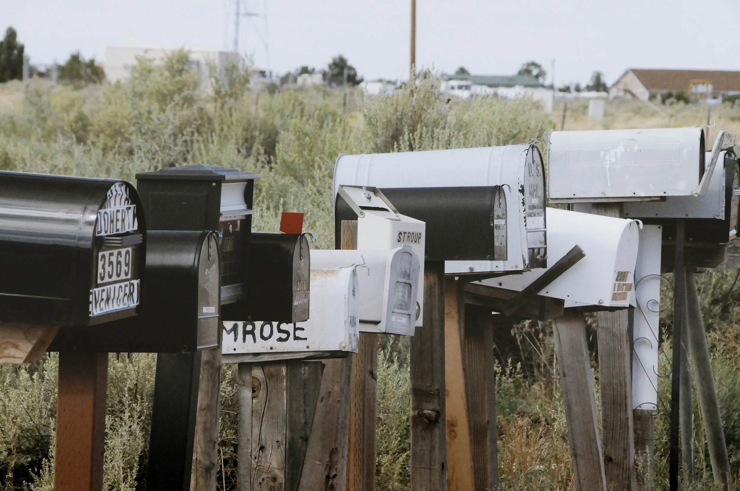 Row of rural mailboxes in various styles and colors along a country road with green foliage in the background.