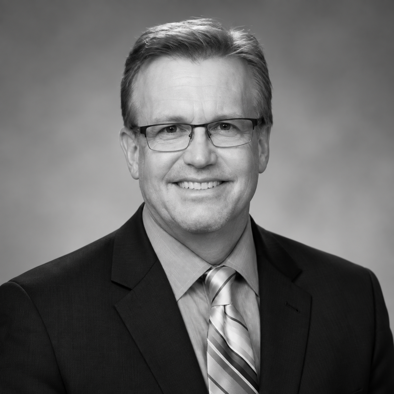 Black and white portrait of a Tim Ecklund wearing glasses, a suit, and a tie, smiling against a plain background.