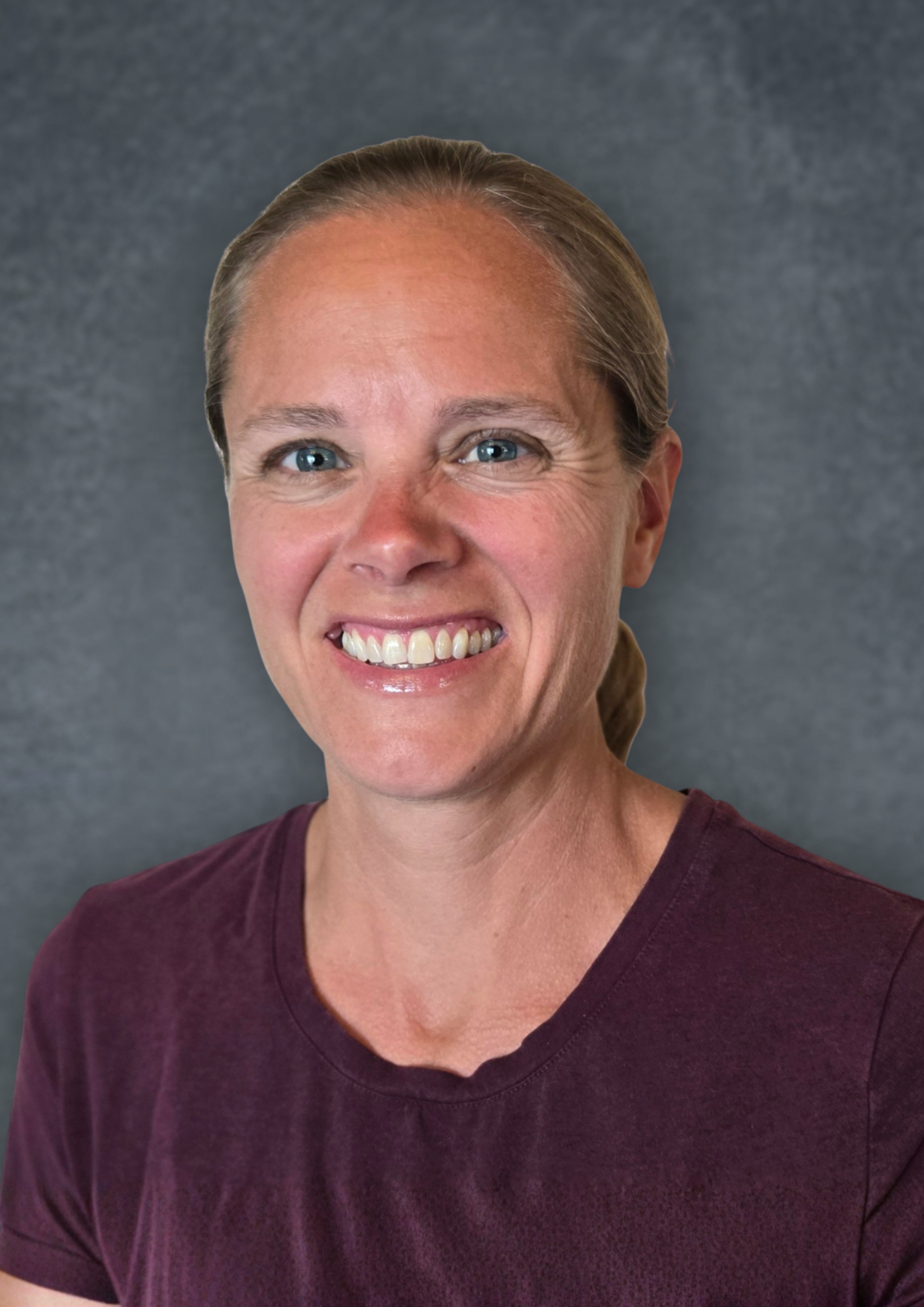 A smiling woman with blue eyes, light skin, and light brown hair pulled back. She is wearing a maroon V-neck shirt and is standing against a textured gray background.