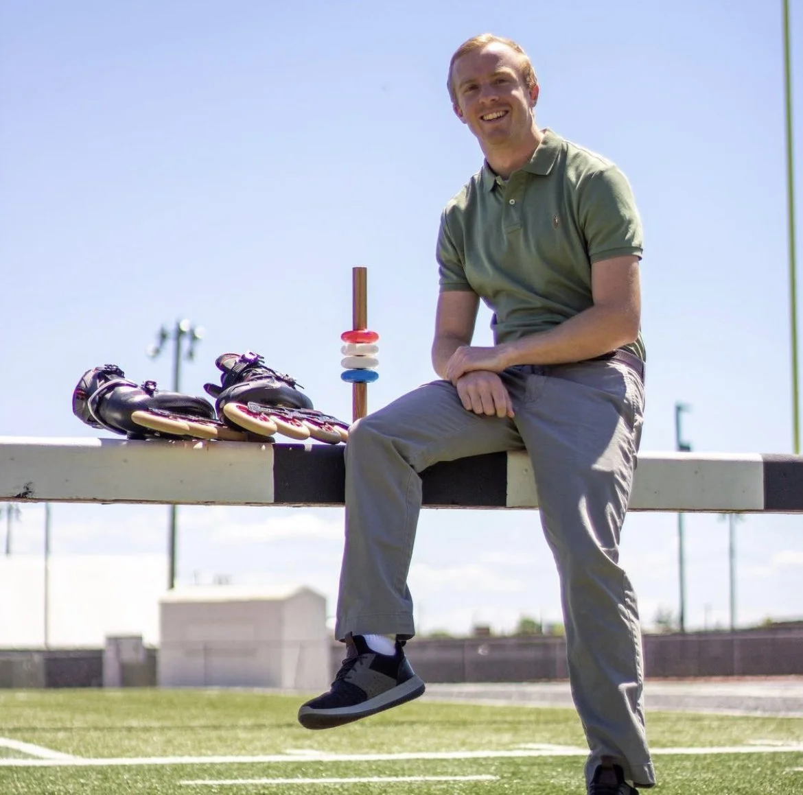 Man sitting on a bench at an outdoor roller skating rink, smiling, with roller skates and a curling broom beside him on the bench.