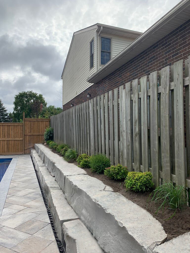 Backyard with a wooden fence, stone retaining wall, and small bushes along a walkway next to a brick house By Logical Landscaping