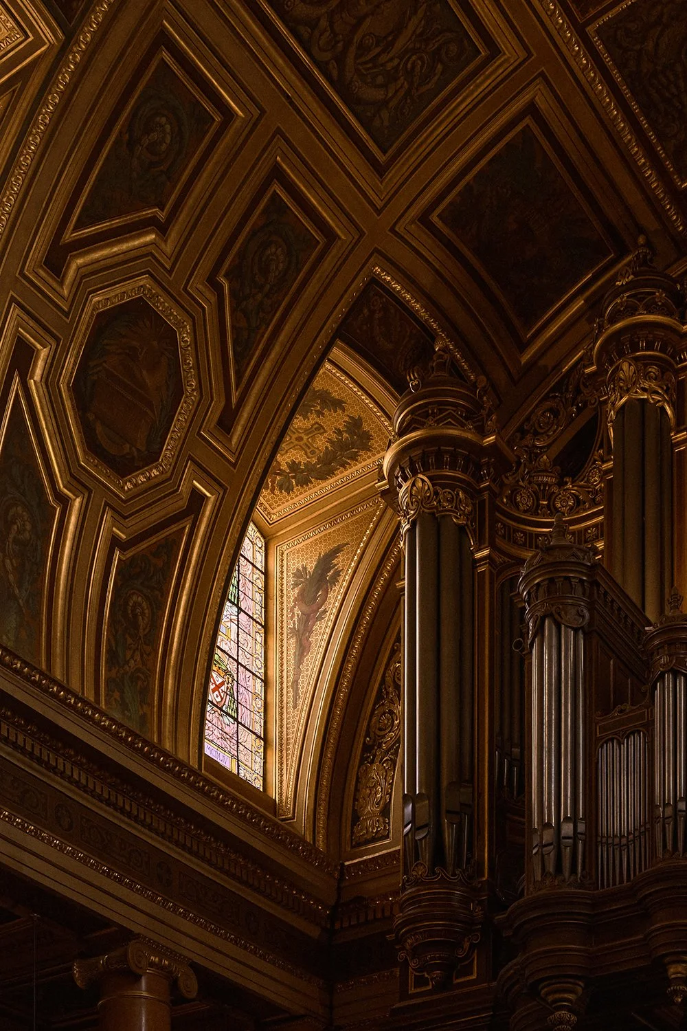 Interior of a grand, ornate church or cathedral with gold accents, detailed woodwork, columns, and a stained glass window.