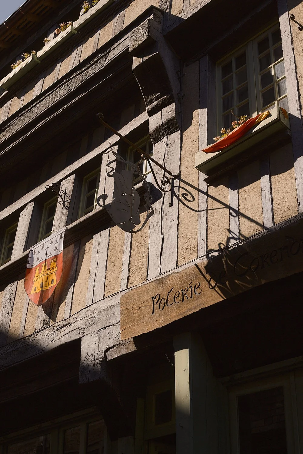Close-up of a historic building with wood and stone exterior, flower boxes on the window ledges, a wooden sign reading 'Poléerie' with French and Polish flags, and a metal sculpture of a woman's face and hair.
