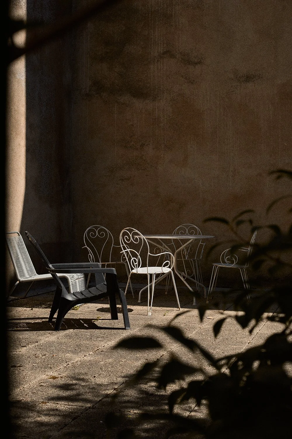 Outdoor patio with black lounge chair, white metal table, and white metal chairs with intricate designs, shadows cast on the ground, against a plain beige wall, partially obscured by dark plant leaves in the foreground.