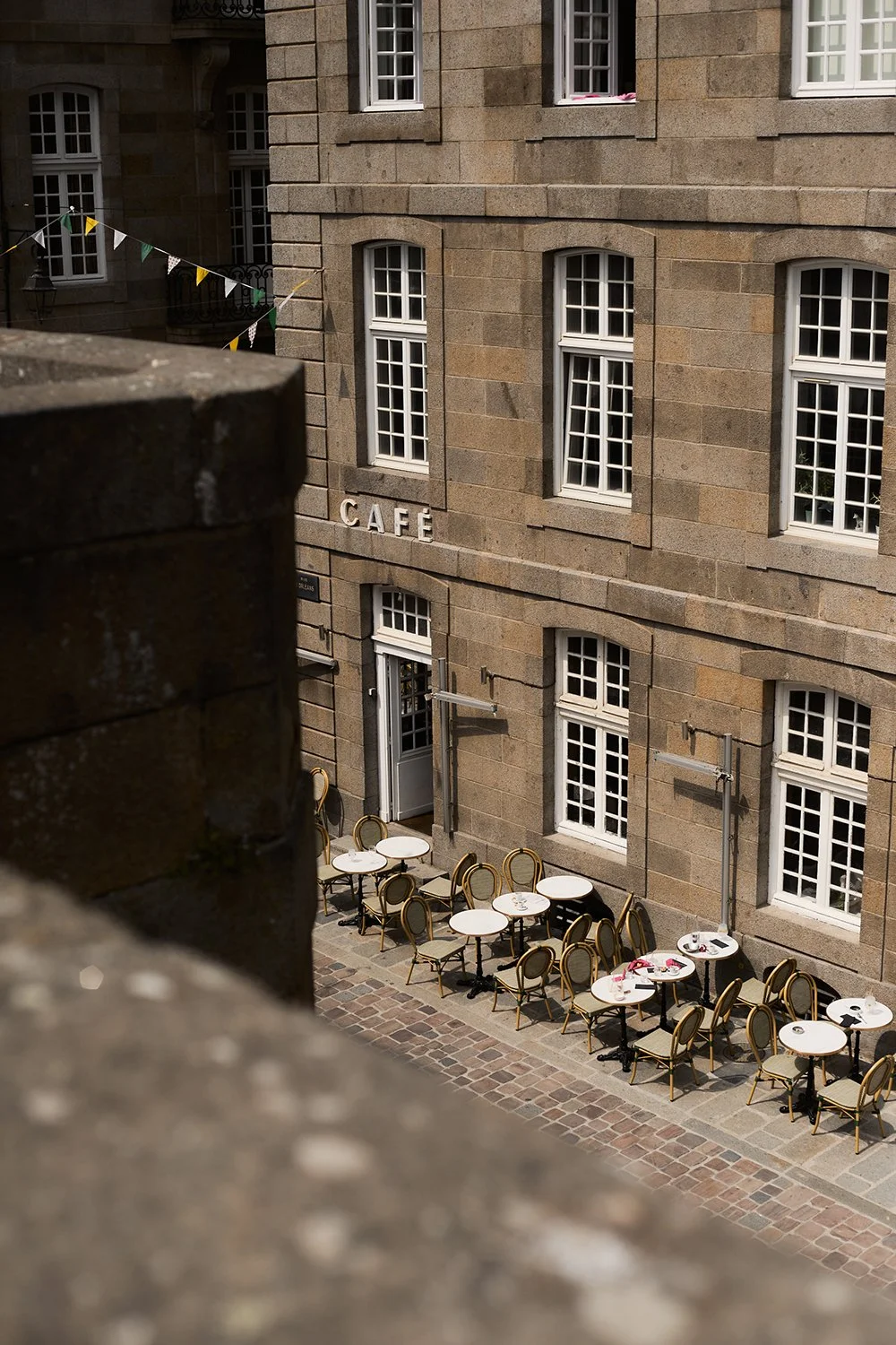 Outdoor cafe with round tables and chairs on a cobblestone street, in front of a stone building with tall windows and a sign that reads 'CAFÉ'.