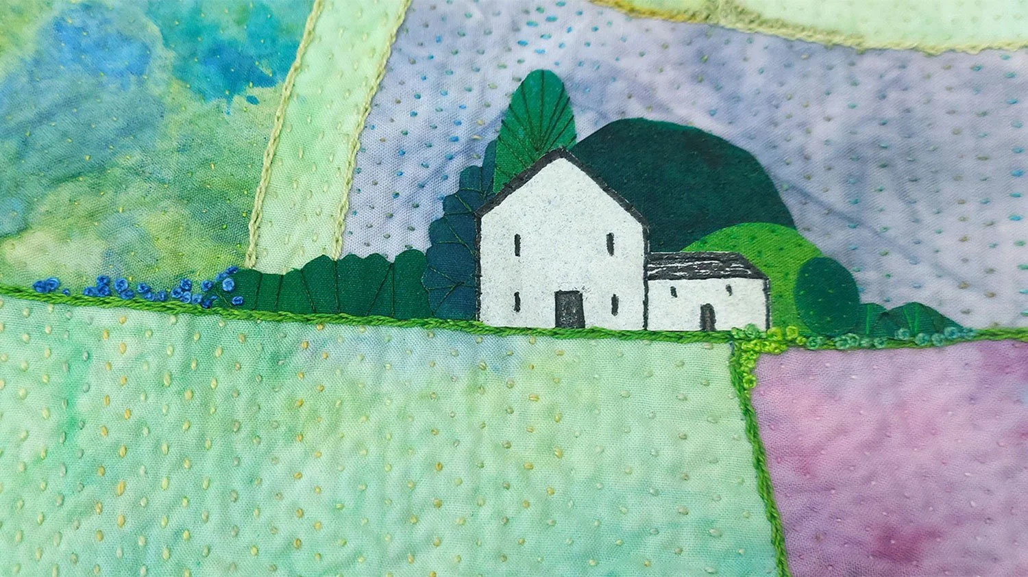 Close-up of fabric with embroidered landscape scene featuring a white house, green trees, hills, and grass, with multicolored sections surrounding it.